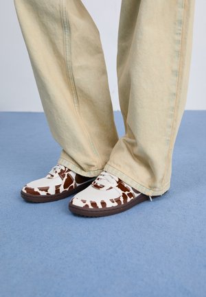 Personne portant un pantalon beige à jambes larges et des baskets blanches avec des motifs de vache marron, debout sur une surface bleue.