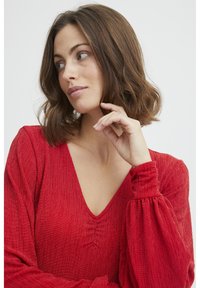 Röd texturerad topp med V-ringning och insynkade detaljer. Långa puffslevar med elastiska smockar. Slätt tyg med subtilt mönster.