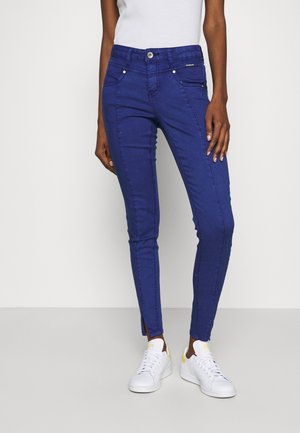 Strakke jeans in levendig blauw denim met een gladde textuur, voorzien van een ritszak, subtiele stikdetails en enkelsplits. Gepaard met witte sneakers.