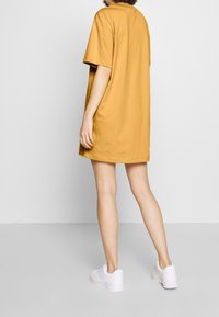 Duża musztardowa sukienka T-shirtowa z bawełny, z okrągłym dekoltem i krótkimi rękawami, zestawiona z białymi trampkami. Prosty, codzienny fason.