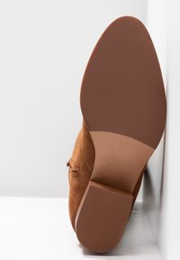 Bottine en daim marron reposant sur son talon, montrant une semelle lisse et un talon texturé sur fond blanc.
