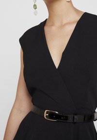 Svart omslagsklänning med korta ärmar, som har en djup V-ringning och ett glänsande svarts patentbälte. Tyget har en texturerad yta.