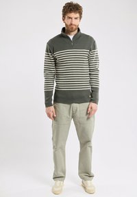 W paskowanym swetrze w ciemnej zieleni i kremowym kolorze. Posiada zamek błyskawiczny przy kołnierzy i ściągacze na rękawach. Połączony z jasnozielonymi spodniami cargo i beżowymi sneakersami.