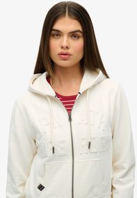 Witte zip-up hoodie met een gestructureerd logo op de voorkant, twee voorzakken, een capuchon met trekkoord en een gelaagd rood-wit gestreept shirt eronder.
