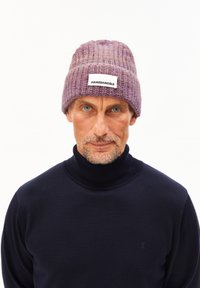 Paarse gebreide beanie met een ribbelstructuur en een platte label met de tekst "ARMEDANGELS." Gecombineerd met een donkere coltrui.