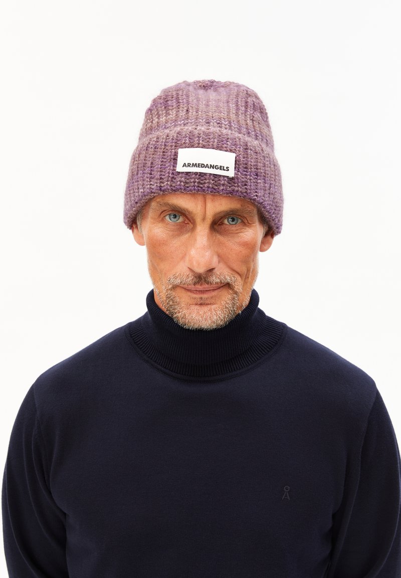 Paarse gebreide beanie met een ribbelstructuur en een platte label met de tekst "ARMEDANGELS." Gecombineerd met een donkere coltrui.
