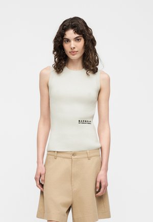 Göndör hajú nő fehér ujjatlan bordázott felsőt és bézs rövidnadrágot visel, „Barbour International” felirattal, egy egyszerű háttér előtt áll.