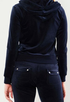 Tuta in velluto blu navy con cappuccio, silhouette aderente e due tasche posteriori, con accenti di bottoni a pressione per un aspetto lineare.