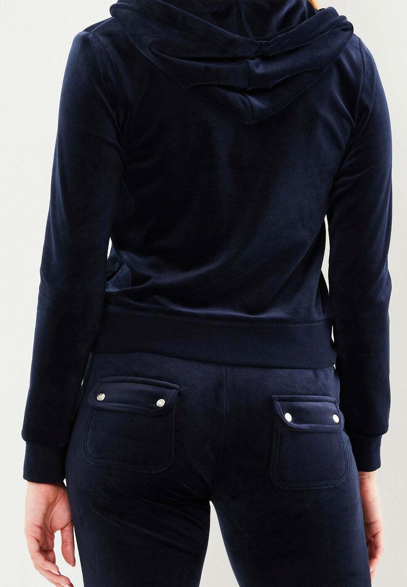 Tuta in velluto blu navy con cappuccio, silhouette aderente e due tasche posteriori, con accenti di bottoni a pressione per un aspetto lineare.