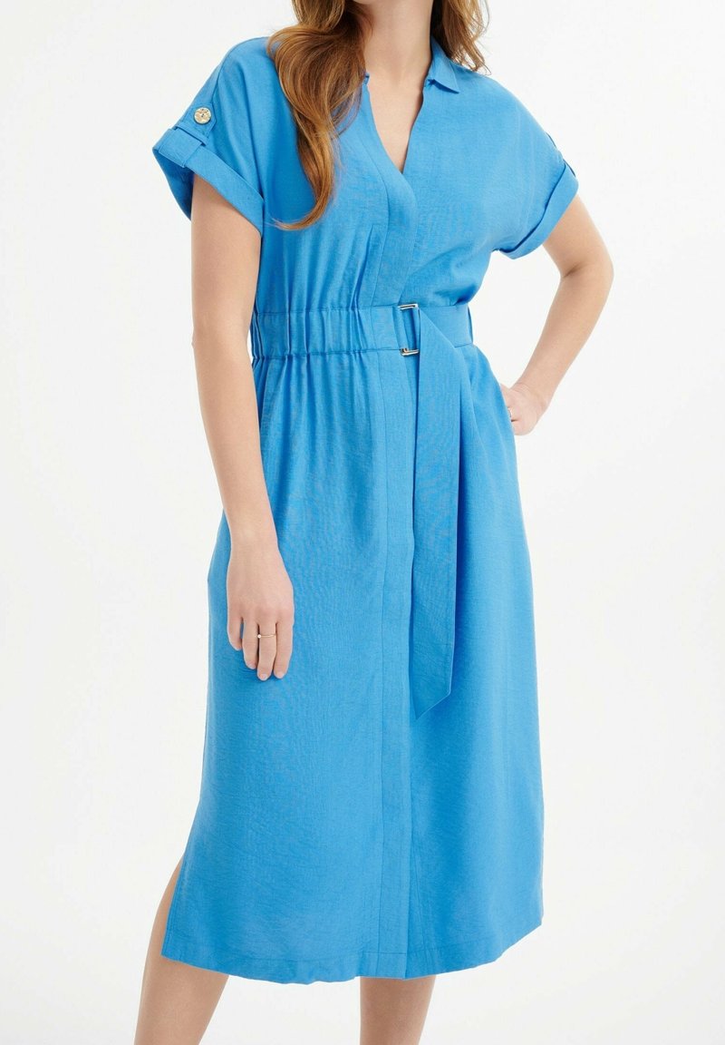 Femme portant une robe bleue à manches courtes arrivant aux genoux avec ceinture et poignet boutonné, posant avec une main sur la hanche devant un fond blanc.