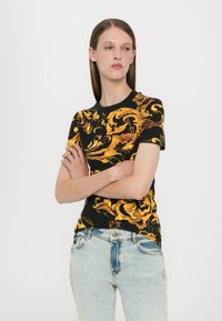 Svart t-skjorte med kort erme som har intrikate gullsurrende mønstre. Rund halsdesign, kombinert med lyseblå denimjeans.