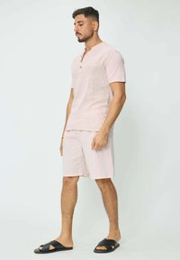 Chemise en lin rose clair à manches courtes avec boutons et col rond, accompagnée d'un short assorti et de sandales noires, sur un fond neutre.