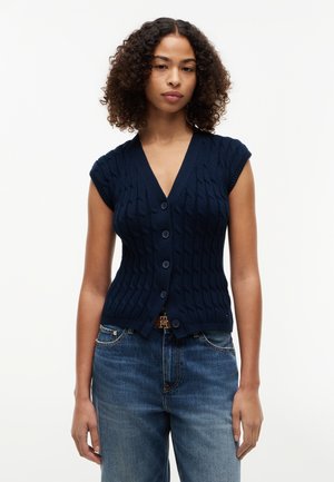 Mladá žena s kučeravými vlasmi, oblečená v tmavomodrom pletenom topu bez rukávov s gombíkmi a modrých džínsoch, stojaca na bielom pozadí.