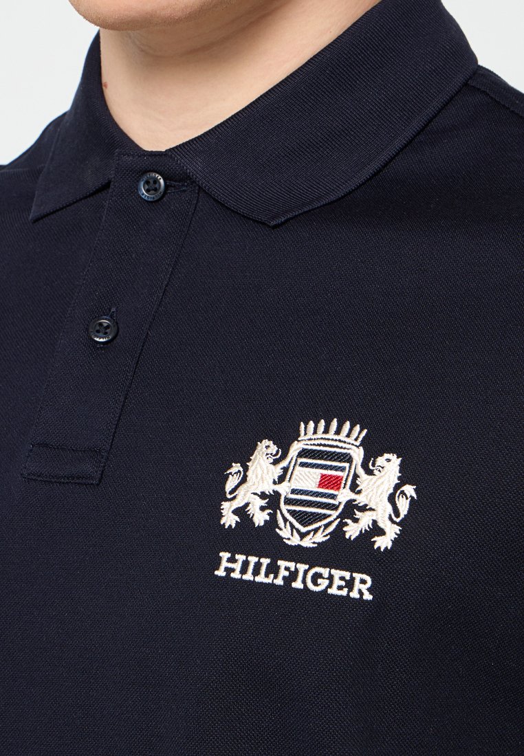 Gros plan sur un polo bleu marine avec deux boutons et un écusson brodé représentant des lions et un drapeau rouge, blanc et bleu au-dessus du texte "HILFIGER".