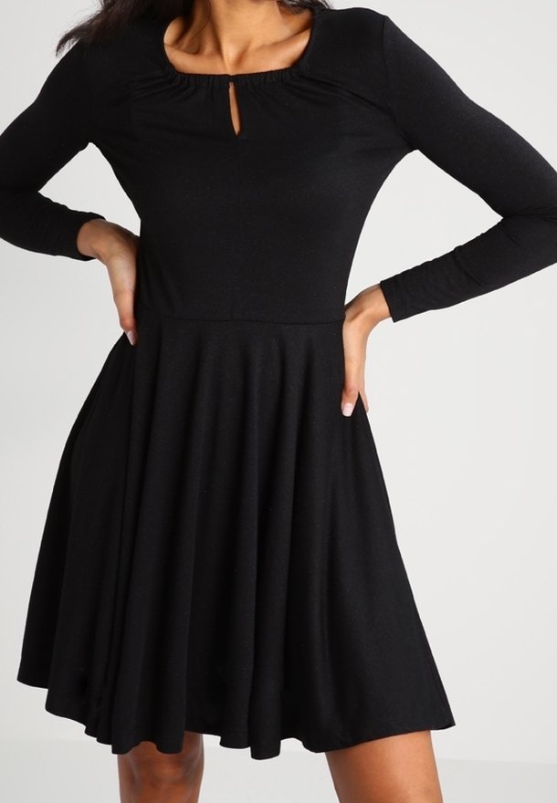 Femme portant une robe noire à manches longues avec une taille ajustée, une jupe évasée et un petit détail d'encolure en forme de clé.