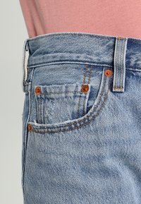Vaqueros de mezclilla azul claro con una textura desgastada, que presentan detalles en cobre, múltiples bolsillos y costuras en contraste en la cinturilla y los bolsillos.