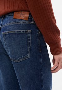 Jeans in denim blu scuro sbiadito con un'etichetta in pelle Calvin Klein sulla cintura. Cuciture visibili delle tasche posteriori e maglione a coste texturato.