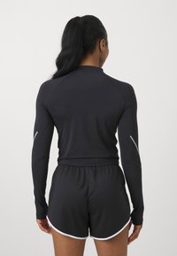 Musta urheilullinen pitkähiha, jossa on raglan-hihat ja heijastavat yksityiskohdat, yhdistettynä mustiin shortseihin, joissa on valkoinen reunus ja joustava vyötärö.