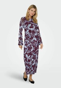 Ilga bordo spalvos suknelė su mėlynais gėlių raštais, ilgomis rankovėmis ir prigludusia forma. Baigta tiesiu apvadu ir dėvima su juodais batais.