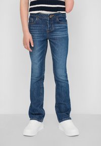 Jean en denim bleu avec une coupe droite, cinq poches et des coutures contrastantes. Porté avec des baskets blanches et un pull rayé.