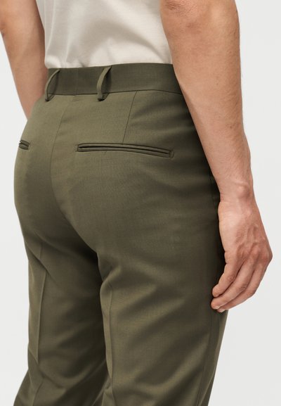 Άνδρας με ελιά πράσινο παντελόνι και λευκό πουκάμισο, εμφανιζόμενος από πίσω, από τη μέση μέχρι το μέσο του μηρού, με το χέρι χαλαρό στο πλάι.