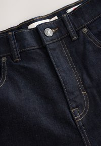 Tmavomodré džínsy z denimu so žltým prešívaním, zapínaním na gombíky a prednými vreckami. Majú štruktúrovanú textúru látky s hladkým zakončením a pevné kovanie.