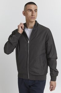 Dark zip-up bomber jacket with ribbed cuffs and hem, featuring side pockets and a smooth, lightweight fabric. Worn over a white t-shirt.