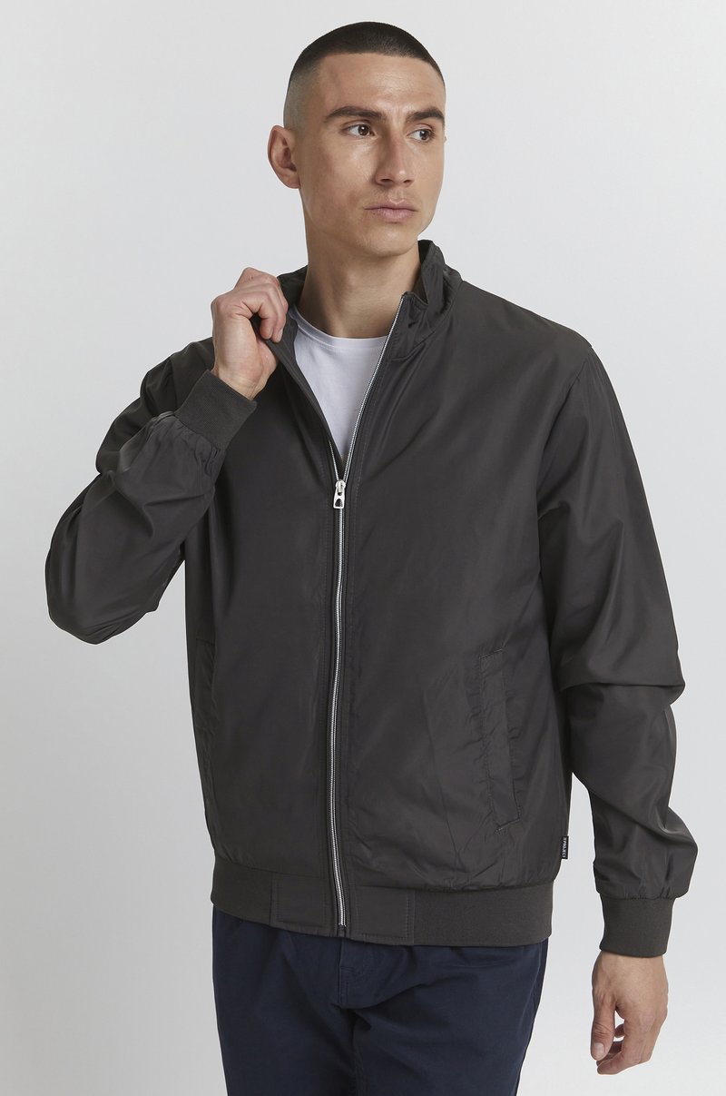 Veste bomber zippée noire avec des poignets et un ourlet côtelés, présentant des poches latérales et un tissu lisse et léger. Portée par-dessus un t-shirt blanc.