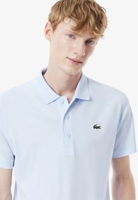 Ljusblå polo skjorta i texturerad tyg, med en två-knapps knäppning och en liten grön krokodil-logotyp på vänster bröst.