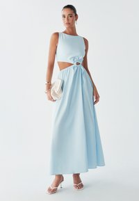 Une femme se tient avec confiance dans une robe maxi bleu clair à découpes, tenant une petite pochette blanche, mettant en valeur des sandales élégantes et des accessoires.