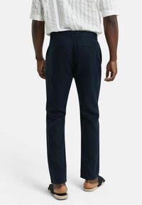 Navy linnen broeken met een rechte pasvorm, voorzien van achterzakken en een subtiele textuur. Gecombineerd met zwarte sandalen en een wit gestreept shirt.