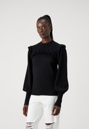 Pull en maille noir avec un empiècement froncé et des manches ballon. Comprend des poignets côtelés et une taille ajustée. Associé à un jean blanc déchiré.