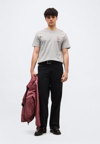 Νεαρός άνδρας με σγουρά σκούρα μαλλιά, φορώντας ανοιχτό γκρι μπλουζάκι με σχέδιο, μαύρο παντελόνι, μαύρα παπούτσια, κρατώντας ένα κόκκινο μπουφάν στο αριστερό του χέρι.