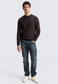 Dunkler Strickpullover mit Rundhalsausschnitt und gerippten Bündchen, kombiniert mit blauen Jeans und weißen Turnschuhen. Legere Passform, strukturiertes Design und kontrastierender Ausschnitt.