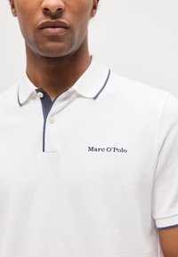 Άνδρας που φοράει λευκό πόλο μπλουζάκι με μπλε σκούρες λεπτομέρειες και το λογότυπο "Marc O’Polo" κεντημένο στο αριστερό στήθος.
