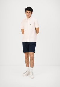 Camisa de botón rosa de manga corta con un bolsillo en el pecho, combinada con pantalones cortos azul marino, calcetines blancos y zapatillas blancas. Diseño sencillo, ajuste casual.