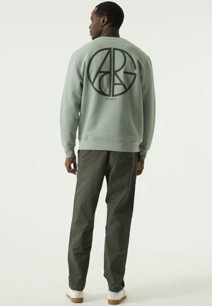 Hombre de espaldas a la cámara, lleva una sudadera verde claro con un gran logotipo circular y pantalones grises, en un escenario de estudio sencillo.