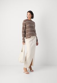 Braun gestrickter Pullover mit weißen Streifen, kombiniert mit einem cremefarbenen Maxirock mit einem Schlitz vorne. Weiße Absatzsandalen und Tasche.