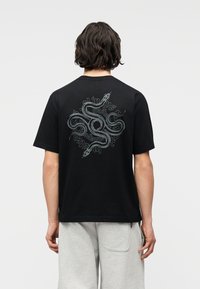 Άτομο με κοντά σκούρα μαλλιά που φοράει μαύρο μπλουζάκι με μεγάλο ασημένιο σχέδιο φιδιού στην πλάτη και ανοιχτό γκρι σορτς.