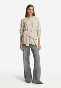 Beige Strickpullover mit einem gebundenen Gürtel-Detail, kombiniert mit grauen weiten Hosen. Der Pullover hat einen runden Halsausschnitt und gerippte Bündchen.