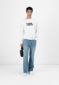 Sudadera blanca con la impresión "KARL LAGERFELD", vaqueros de campana azules, zapatillas blancas, sosteniendo un bolso negro, de pie contra un fondo liso.