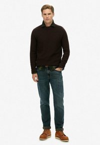 Bruine gebreide trui boven een groen geruit hemd, gecombineerd met blauwe spijkerbroek. Bruine leren schoenen met veters en een zichtbare horloge.