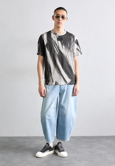 Fekete-fehér grafikai póló absztrakt ecsetvonás mintával, világoskék, laza szabású farmerral és fekete sportcipővel párosítva. Szürke háttér.