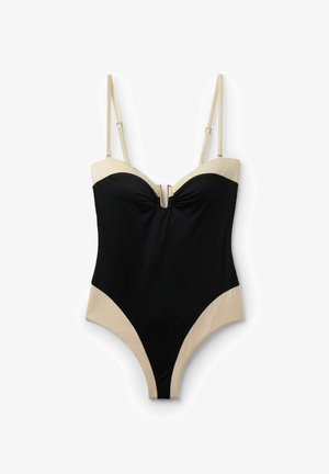 Schwarz-beiger Einteiler mit verstellbaren dünnen Trägern und einem goldfarbenen Reißverschlussdetail vorne in der Mitte der Brust.