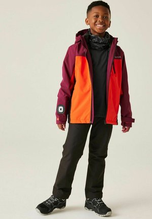 Young boy smiling, wearing a color-block orange and maroon jacket, black pants, black shoes, and a patterned neck gaiter, standing against a plain background.