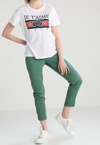 T-shirt blanc avec un texte graphique noir et rouge et un imprimé de rose, associé à un pantalon vert cropped et des sneakers blanches avec des lacets noués.