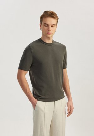 Suéter corto de manga corta en verde oscuro, fabricado en un material de punto ligero, con un escote redondo y bordes acanalados. El modelo lleva unos pantalones de color claro.