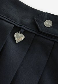 Dunkelblaßer, gefalteter Stoff mit einem silbernen, herzförmigen Anhänger, der den Markennamen "Clarks" trägt, sowie einem silbernen Knopfakzent am Bund.