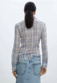 Lightweight, plaid button-up shirt with long sleeves in shades of blue and gray, featuring a curved hem and cuff details, paired with a denim skirt.