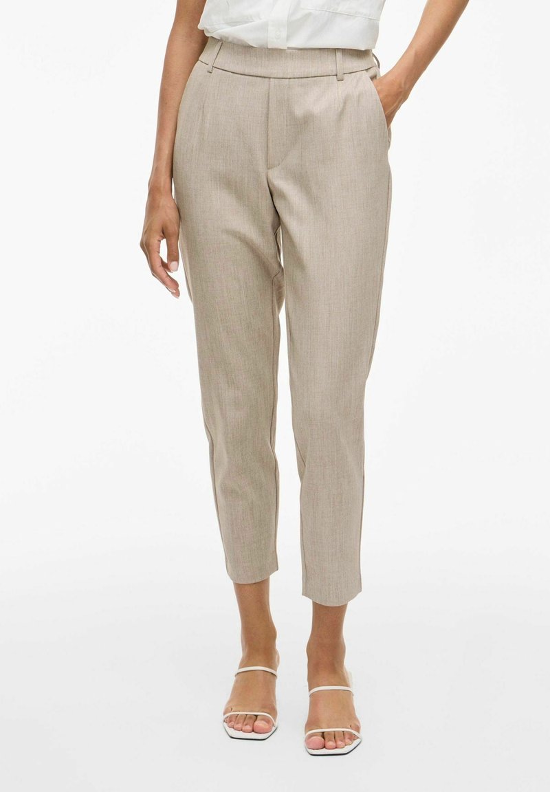 Femme portant un pantalon beige taille haute fuselé avec poches, associé à des sandales blanches à brides ouvertes à l’avant et un chemisier blanc.